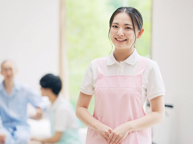 笑顔のスタッフが立つ居室 居室 自然光が差し込む明るい空間で、スタッフが笑顔で利用者を迎えています。アットホームな雰囲気を感じられます。