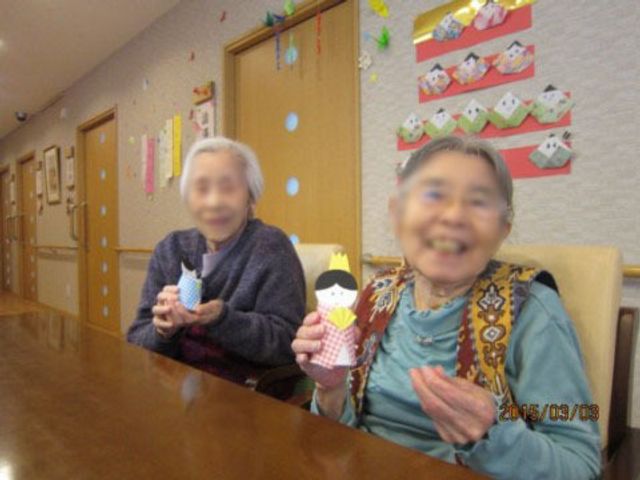 楽しげな高齢者二人の居室 ご入居者様 笑顔で手芸品を持つ二人の高齢者がいる居室です。ここではご入居者が趣味の時間を楽しむ様子が伺えます。