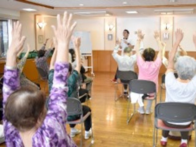 高齢者の運動風景 機能回復訓練 指導員による運動プログラムが行われており、参加者は元気に手を挙げている様子です。明るく広々とした空間で、活動的な日々を送ることができます。