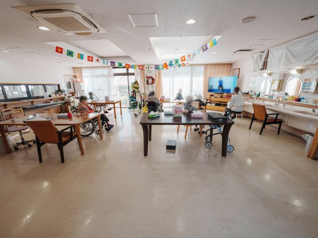 明るい共有スペース ラウンジ 広々とした明るいスペースで、入居者の方々がゆったりとリラックスできる環境です。人々が椅子に座り、テレビを楽しむ様子があります。