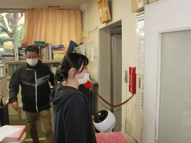 職員がいるエントランス エントランス   受付まわりで職員の方が電話対応をしている様子です。書棚や掲示物があり、落ち着いた雰囲気が伝わります。