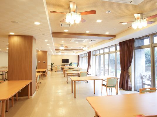施設の写真 広い食堂は温かみのあるデザインである。壁にはテレビが設置されている。片側は一面窓になっているので、外の光が入り、とても明るい。