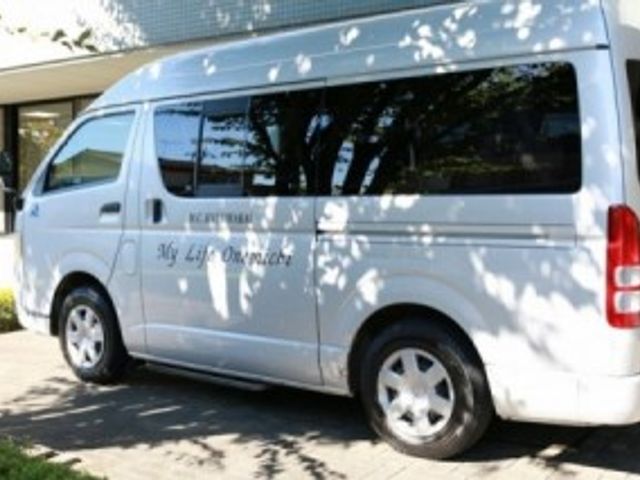 ケアを感じる送迎車 エントランス 木漏れ日が映る送迎車は、お出かけや通院の際の心強い味方です。安心のドライブをサポートします。