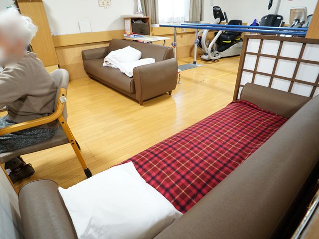 居室のベッドとソファ 共有スペース 広々とした木目調の床が印象的な居室です。ゆったりしたソファが配置され、くつろぐ人の姿が見えます。