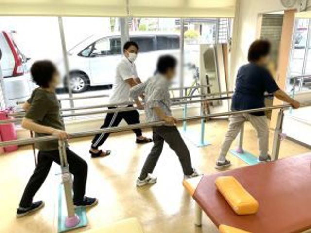 運動を行う人々と職員の機能訓練室 機能訓練室   自然光がたっぷり入る中で、職員が利用者の運動をサポートしています。サポート体制が整っています。