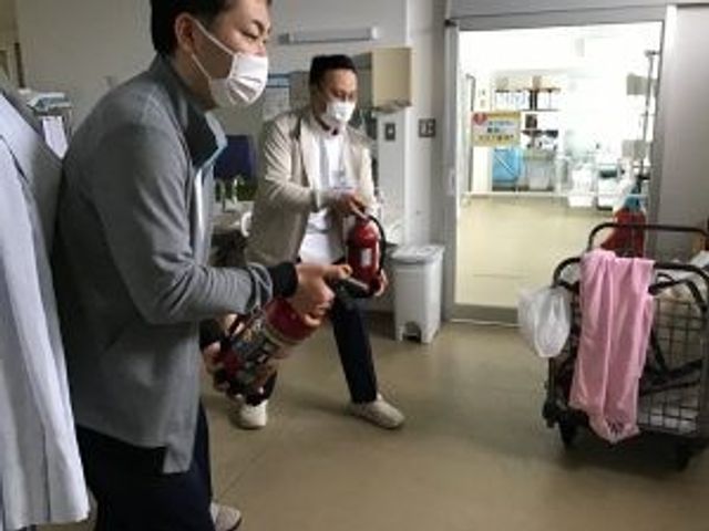 消火訓練の廊下 廊下   職員の方が消火器を持って確認している、落ち着いた雰囲気の廊下です。実践的な訓練の場面が見られ、安心につながる取り組みがうかがえます。
