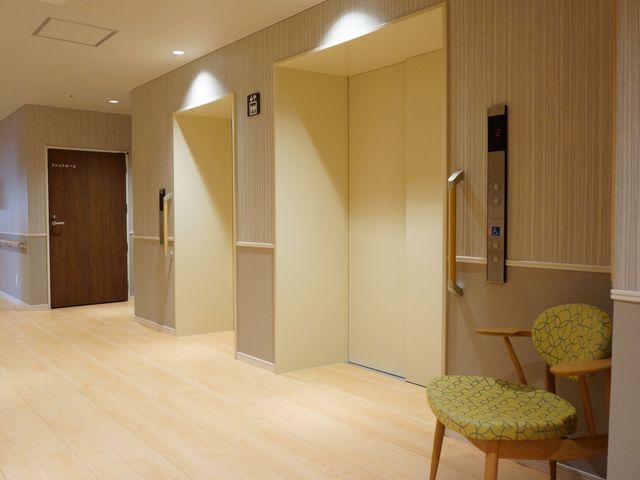 エレベーターと椅子の廊下 エレベーター ゆったりとした廊下には木目調の椅子が置かれ、待ち時間も心地よく過ごせる空間が演出されています。明るく清潔感のあるデザインで気持ちも穏やかになります。