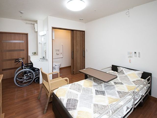 車椅子のある居室 居室   木目調の床と白い壁で、落ち着いた雰囲気の居室です。ベッドのそばに椅子があり、出入りしやすい配置に見えます。