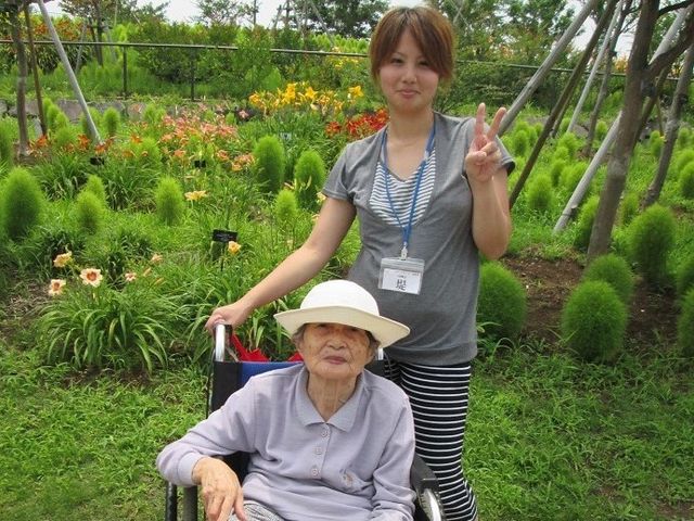 庭園で笑顔のスタッフと利用者 外出 緑あふれる庭園で、スタッフが利用者様のそばで明るくサポートしており、二人の笑顔が印象的です。快適な自然環境も魅力のひとつです。