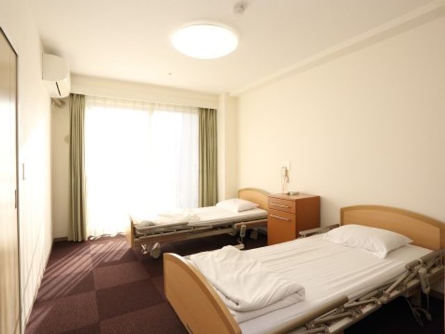 清潔なベッドの居室 ２人部屋 ゆったりとしたベッドと清掃が行き届いた室内です。大きな窓で明るく、落ち着いた時間を過ごせる環境が整っています。