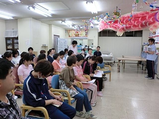 説明を聞く利用者の集まり 多目的室   広い多目的室で、利用者の方々が椅子に座って説明を聞いています。天井が高く、行事にも使いやすそうな空間です。