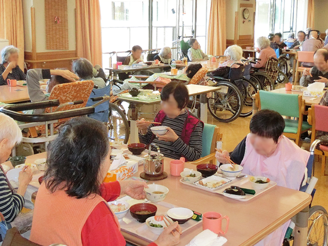 食事を楽しむ人々食堂 食堂   自然光が差し込む明るい空間で、多くの人々が楽しそうに食事をしています。車椅子利用の方も多く見られます。