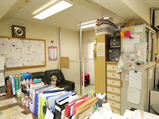 施設の写真 テーブルの上にはファイルや書類が多く並べられている。壁に掛けられたホワイトボードにも、書類が埋め尽くされている。