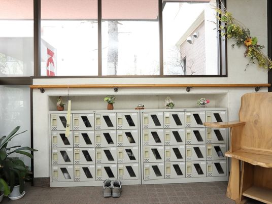 施設の写真 下駄箱の横には木のベンチが用意されている。下駄箱の上には花が４つ飾ってある。手すりに靴ベラが引っ掛けてある。