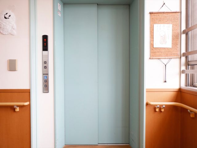 青みがかったエレベーター扉 エレベーター 手すりが付いた壁と、明るい色のエレベーターが、快適な移動をサポートします。安全に配慮した構造で、ご高齢の方も安心です。