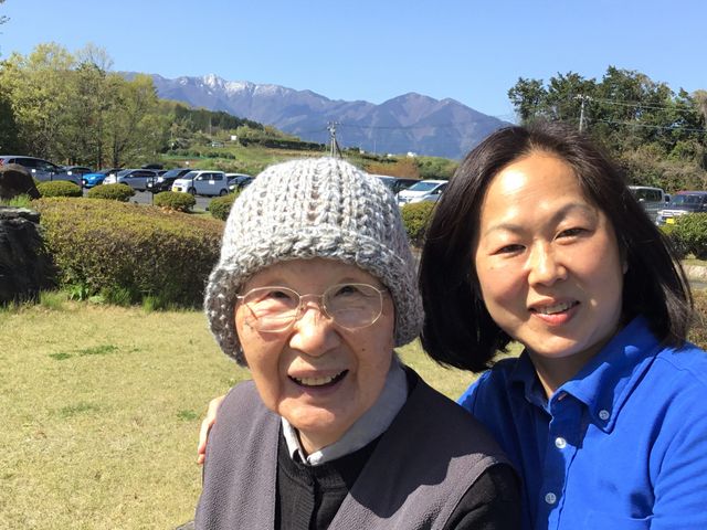 笑顔の二人中庭にて 中庭で穏やかなひとときを過ごす高齢者とスタッフ。背景には山々が広がり、開放的な環境が感じられます。
