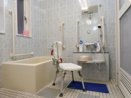 グレー色のタイル調の模様入りの壁。浴槽の隣に洗い場が設置。正面には鏡があり、その下部は台になっている。滑り止めの付いた椅子が置かれている。