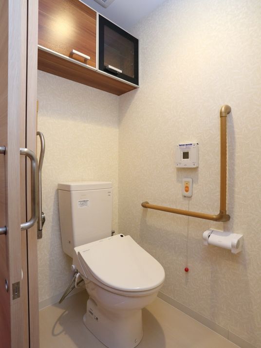 居室内のトイレには、温水洗浄便座がある。便座の横には手すりがある。手に届きやすい位置にナースコールが設置されている。