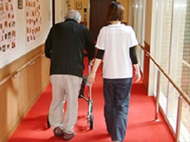 歩行補助を受ける高齢者 通路 手すり付きの明るい廊下で、スタッフが優しくサポートしながら、入居者が安心して歩行訓練を行っています。