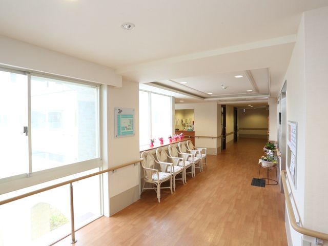 廊下にある椅子と掲示物 廊下 明るく開放的な廊下は、ゆったりとした椅子が配置され落ち着いた雰囲気を醸し出しており、ちょっとした休憩にも適しています。掲示された情報は日々の生活に役立つ内容でしょう。