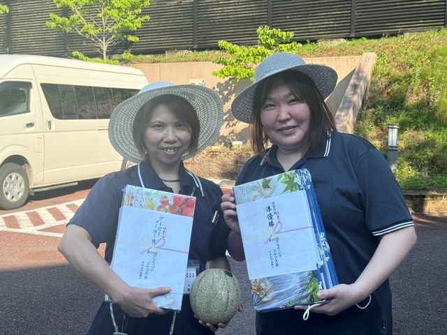 屋外で賞品を持つ職員 スタッフイメージ   屋外で笑顔の職員お二人が、賞状のようなものとメロンを手にしています。明るい雰囲気が伝わり、働く人の和やかな様子が見られます。