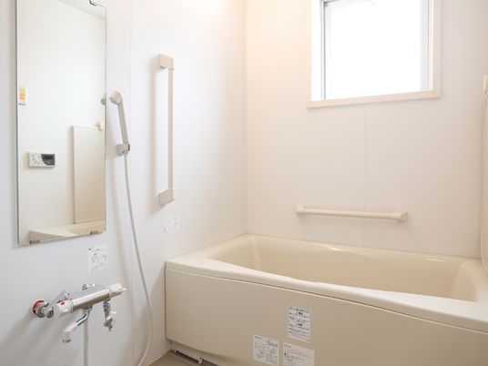 浴室は、全体的に白でまとめられたデザインになっている。窓から明るい光が差し込んでいる。壁には大きな鏡や手すりがある。