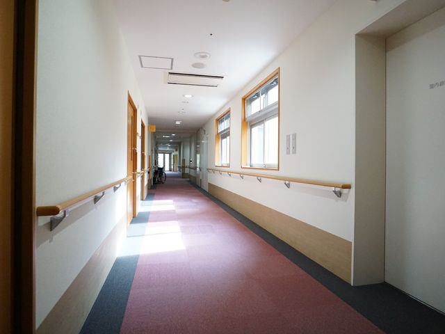 明るい廊下の様子 廊下 手すりが設けられた安全に配慮した廊下です。奥には車椅子が見え、バリアフリー環境が整っていることが伺えます。