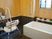 サムネイル 浴室には壁面にハンドシャワーと鏡が取り付けられ、前に椅子が１脚置いてある。小窓側の壁際には浴槽が設置され、脇に手すりと洗面器が並んでいる。