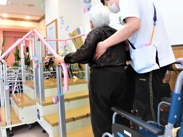 機能訓練室でサポート 機能訓練室 スタッフの方がサポートしながら、バーをつかみ歩行訓練を行っている様子です。明るい雰囲気の中、リハビリに励んでいます。