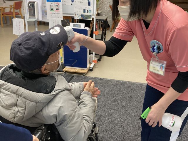 受付での体温測定シーン フロント 施設の玄関では丁寧に体温測定が行われ、スタッフが利用者をサポートします。安心して過ごせます。