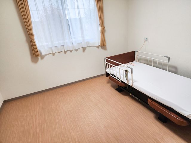 シンプルな居室のベッド 入居者様 ゆったりとした床空間が特徴の居室です。清潔感のある寝具と窓からの自然光が心地よい空間を作り出しています。
