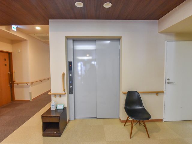 廊下のエレベーター前 エレベーター 手すりが設置された廊下に、座面のある待合椅子が備えられており、快適に移動のお待ちいただけます。安全かつ円滑な移動を支援する環境が整っています。