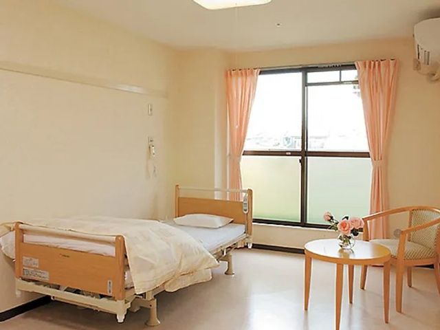 明るい居室のベッド 居室   大きな窓から光が入る、落ち着いた雰囲気の個室です。ベッドのそばにテーブルと椅子があり、くつろぎやすい空間です。