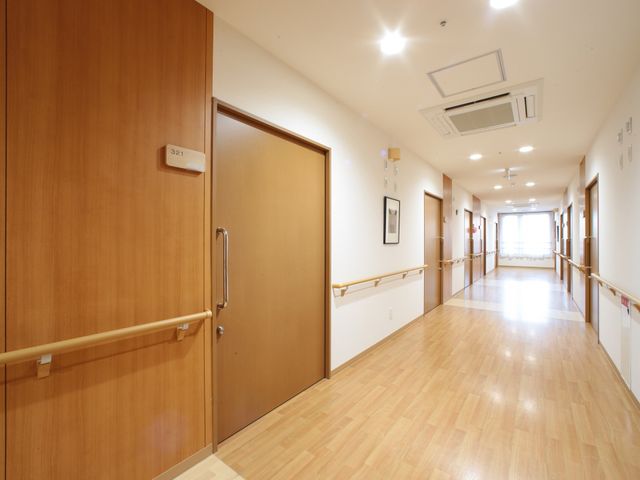 明るい廊下と居室の扉 通路 すっきりとした木目調の廊下が広がり、安全に配慮された手すりが設置されています。白壁とのコントラストが居心地の良さを感じさせます。