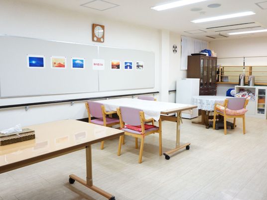 壁には手すり、富士山の写真が飾られている。壁掛け時計もある。奥には茶箪笥、電気ポットが置かれている。部屋全体にはテーブルと椅子がある。