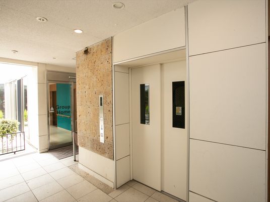 施設館内に完備されている白い扉のエレベーターである。広々としたホールで、左側には玄関の自動扉が見える。