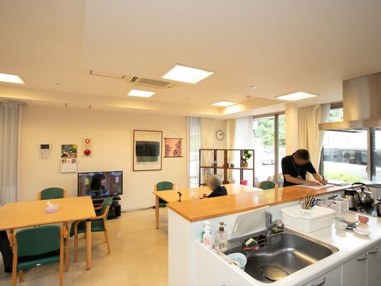 食堂に備わっているキッチンカウンターで男性が作業している。その後ろに、テレビを視聴している入居者様の姿が見える。