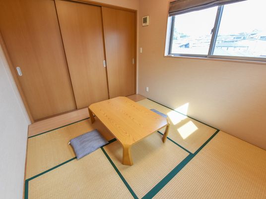 畳の敷かれた和室には、中央に机が一つと座布団が設けられている。奥には収納スペースがある。窓の外には住宅地が広がっている。