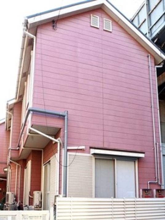 三角屋根にピンクの外壁の施設は2階建てで、一般家庭のような外観になっている。雨戸や塀は白くなっている。