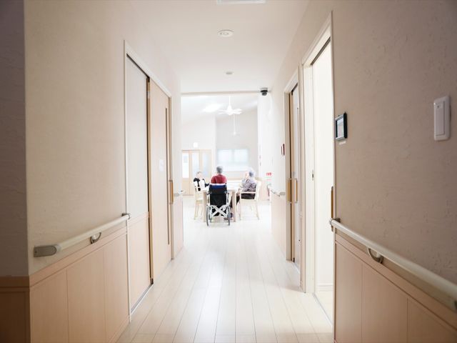 廊下と居室の風景 廊下 清潔感のあふれる廊下が続き、先にはゆったりと談笑する人々の姿が見えます。落ち着いた雰囲気の中でのびのびとお仕事できる環境です。