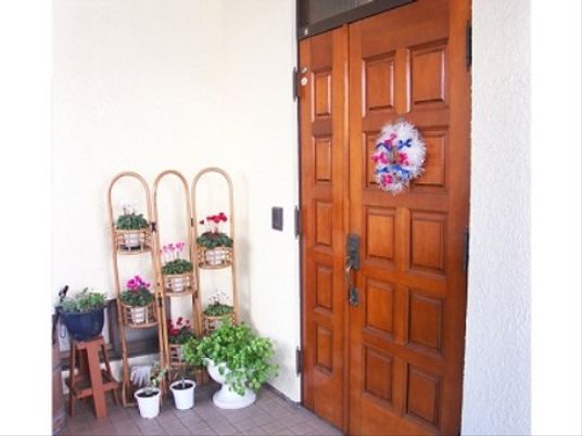 玄関ドアには、白に青と赤のリボンを付けたリースを掛けている。横側に花やグリーンの鉢を台や地面に置いて飾っている。