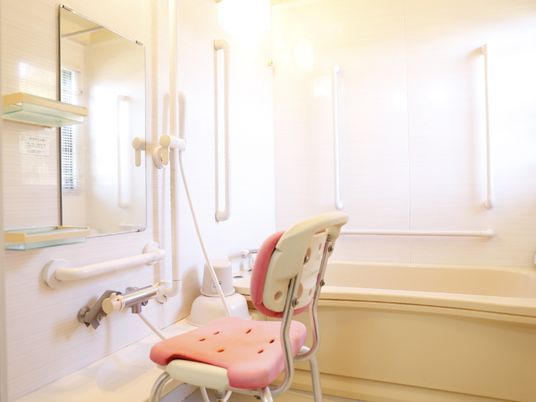 白を基調としたバスルームである。洗い場には座面と背もたれがピンク色の椅子が置かれている。壁には手すりがある。