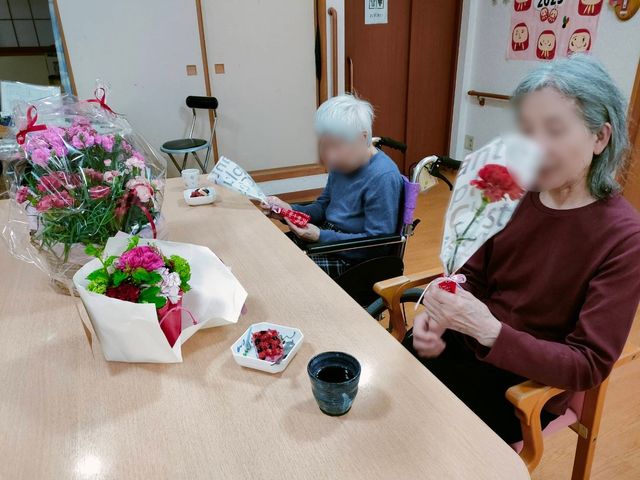 高齢者が花を持つ様子 行事 笑顔で華やかな花を手にされた高齢者の方々が、温かみのある食堂で心地よい時間を過ごされています。