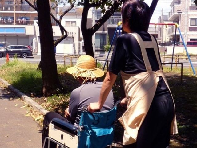 外でくつろぐ高齢者と介護者 散歩 ゆとりある中庭で日差しを楽しむ高齢者とスタッフの姿です。自然に囲まれた空間での安らぎの時間をサポートしています。