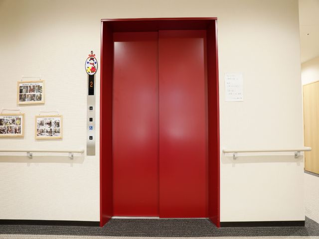 赤い扉のエレベーター前 エレベーター 広々とした廊下に面した赤いエレベーターは、利用者の移動を快適にサポートします。壁面には活動の様子が掲示され、温もりが感じられる空間です。