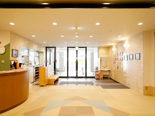 明るいエントランスの様子 玄関 温かみのあるデザインのエントランスで、スタッフの笑顔がお出迎えします。ジャケットを着用した方が笑顔です。