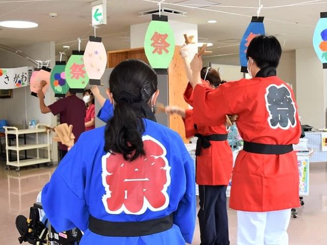 盆踊りを楽しむ人々の背中 多目的室 彩り豊かな飾りつけが施された室内で、和服姿の方々が盆踊りを楽しんでいる様子です。お互いの交流も深まる楽しいイベントが行われています。