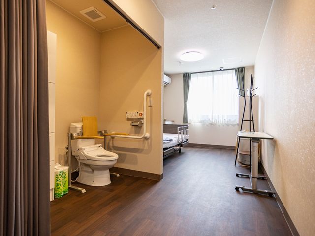 居室のトイレとベッド 居室   手すり付きのトイレがあり、移動しやすさに配慮されたつくりです。窓から光が入る、落ち着いた居室です。