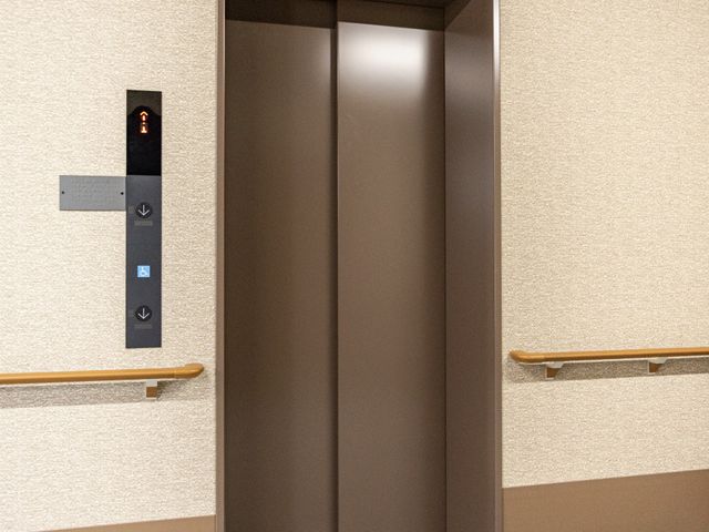 エレベーター前の廊下 廊下   手すりが設置された落ち着いた廊下に、エレベーターの出入口があります。床には誘導用の点字ブロックがあり、移動しやすい工夫が見られます。