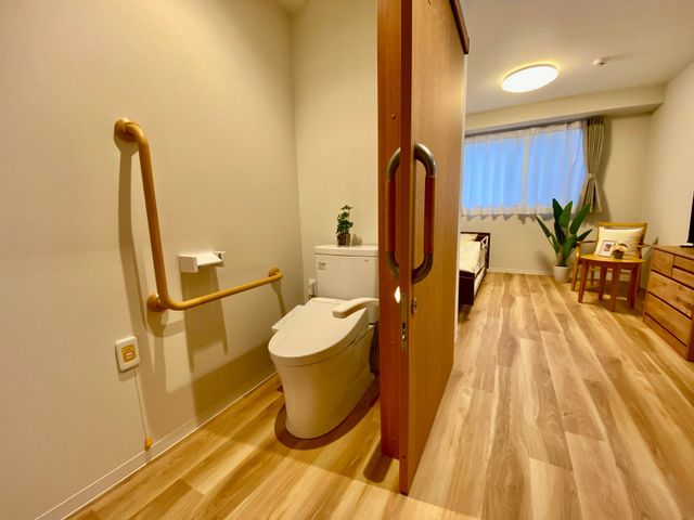 手すり付き居室トイレ 居室   手すりが付いたトイレと、木目調で明るい居室が並ぶ落ち着いた空間です。窓から光が入り、家具もすっきり配置されています。